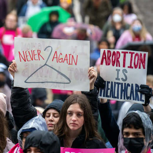 two humans holding signs as protest that read "never again" with a hanger and "abortion is healthcare"