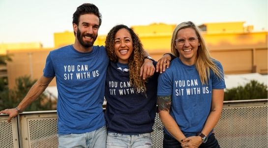 photo of 3 humans smiling at the camera with only human shirts that read "you can sit with us"